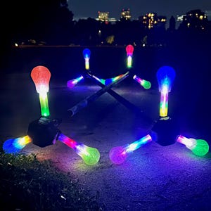 May include: A nighttime shot of illuminated outdoor lights. The lights are arranged in a starburst pattern, with each light emitting a vibrant spectrum of colors. The background features a dark park setting with city lights in the distance.