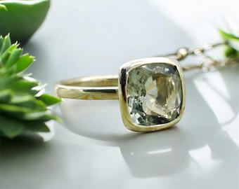 May include: A gold ring with a large, square-shaped, light green gemstone. The ring is set in a simple, modern style.