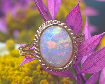 May include: A gold ring with an oval-shaped opal gemstone. The opal is iridescent with shades of pink, green, and blue. The ring is set in a delicate, rope-like band.