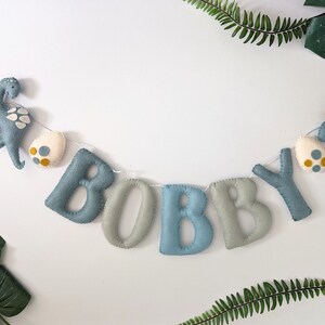 Pode incluir: Uma faixa de feltro com o nome "BOBBY" escrito em letras azuis e cinzas. A faixa &eacute; decorada com dois dinossauros de feltro e ovos.