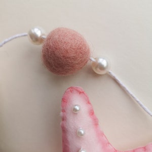 May include: A pink felt starfish ornament featuring white stitched edges and small white pearl beads attached to its surface. Above the starfish, a white string holds a single round dusty pink felt ball flanked by two white pearl beads.