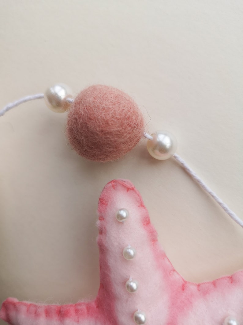 May include: A pink felt starfish ornament featuring white stitched edges and small white pearl beads attached to its surface. Above the starfish, a white string holds a single round dusty pink felt ball flanked by two white pearl beads.