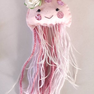 May include: A pink felt jellyfish with a white flower on top. The jellyfish has pink glitter accents and white and pink yarn tentacles.