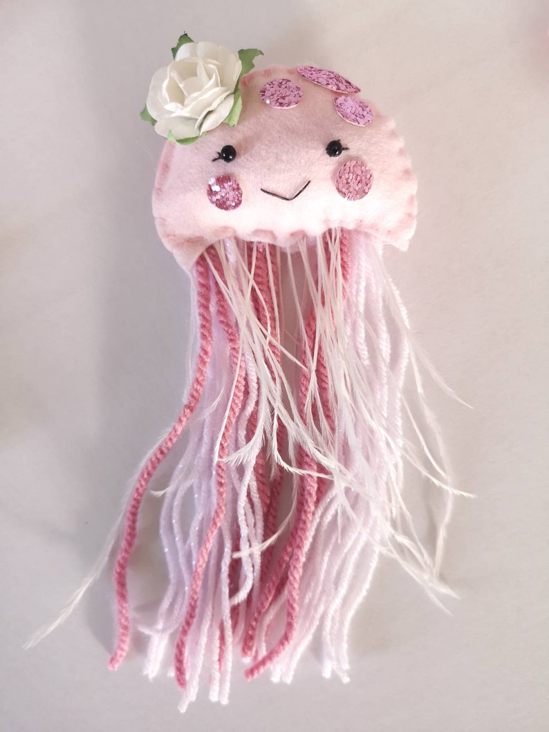 May include: A pink felt jellyfish with a white flower on top. The jellyfish has pink glitter accents and white and pink yarn tentacles.