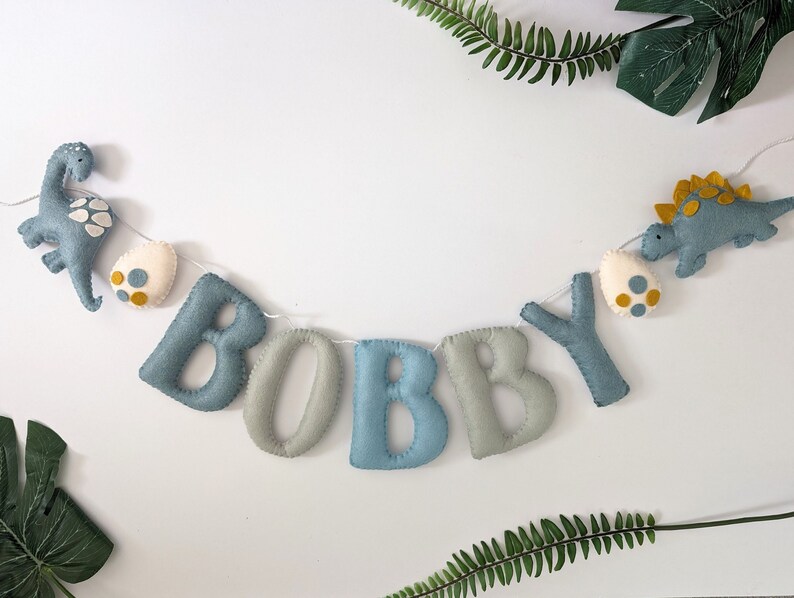Pode incluir: Uma faixa de feltro com o nome "BOBBY" escrito em letras azuis e cinzas. A faixa &eacute; decorada com dois dinossauros de feltro e ovos.