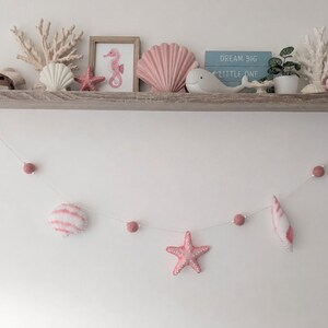 May include: A wooden shelf displays nautical decor including white coral, a seahorse print, a pink shell, a white whale, a small plant, and a lighthouse. Below, a string garland features pink felt balls, a soft shell, and a pink starfish.
