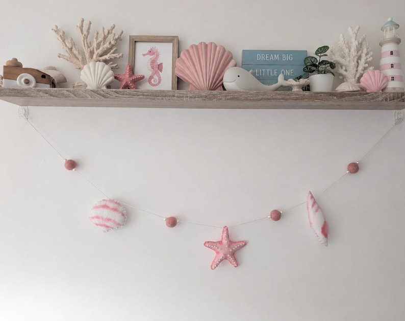 May include: A wooden shelf displays nautical decor including white coral, a seahorse print, a pink shell, a white whale, a small plant, and a lighthouse. Below, a string garland features pink felt balls, a soft shell, and a pink starfish.