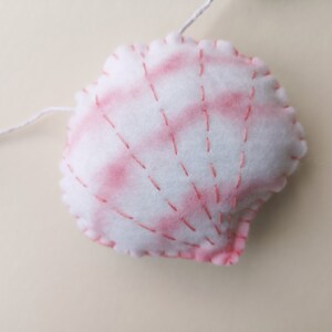 May include: A white felt seashell ornament featuring soft pink watercolor-style shading and decorative pink stitched lines radiating from the base. The plush shell is attached to a thin white string, showcasing hand-stitched edges in pink thread.