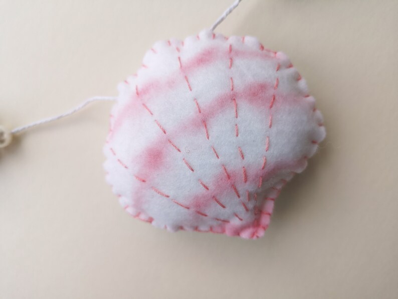 May include: A white felt seashell ornament featuring soft pink watercolor-style shading and decorative pink stitched lines radiating from the base. The plush shell is attached to a thin white string, showcasing hand-stitched edges in pink thread.