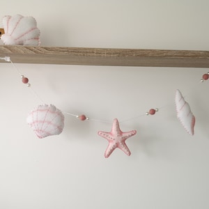 May include: A decorative garland featuring white and pink felt sea creatures including a scallop shell, a starfish, and a conch shell. The items are strung together on a thin white cord interspersed with small white beads and dusty pink felt balls.