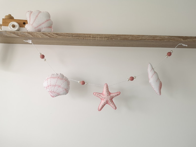 May include: A decorative garland featuring white and pink felt sea creatures including a scallop shell, a starfish, and a conch shell. The items are strung together on a thin white cord interspersed with small white beads and dusty pink felt balls.