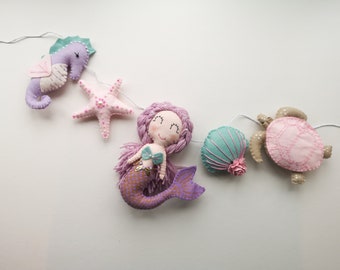 May include: A felt garland with a mermaid, a starfish, a seashell, a seahorse, and a turtle. The garland is strung on a white string and is perfect for a nursery or child's room.