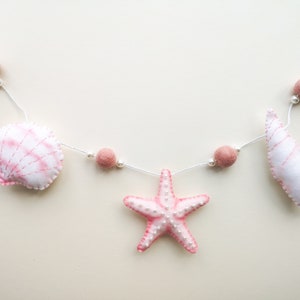 May include: A decorative garland featuring plush fabric shapes of a seashell, a starfish, and a spiral shell in shades of white and soft pink. The items are strung on a white cord interspersed with small white beads and dusty rose felt balls.