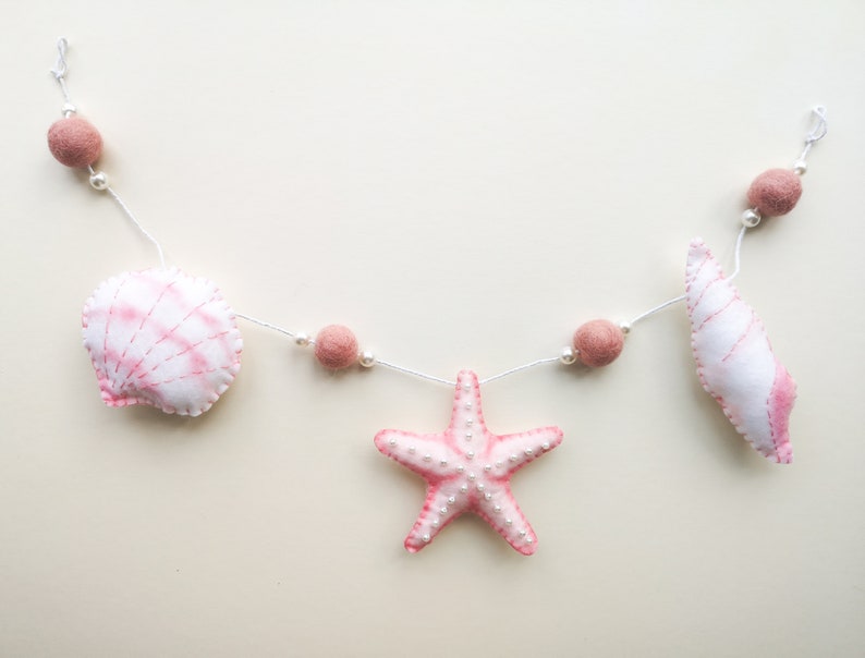 May include: A decorative garland featuring plush fabric shapes of a seashell, a starfish, and a spiral shell in shades of white and soft pink. The items are strung on a white cord interspersed with small white beads and dusty rose felt balls.