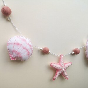 May include: A decorative garland featuring felted shapes including a white and pink scallop shell, a pink starfish, and a smaller white and pink shell. The shapes are strung on a white cord with dusty rose felt balls and small white beads.