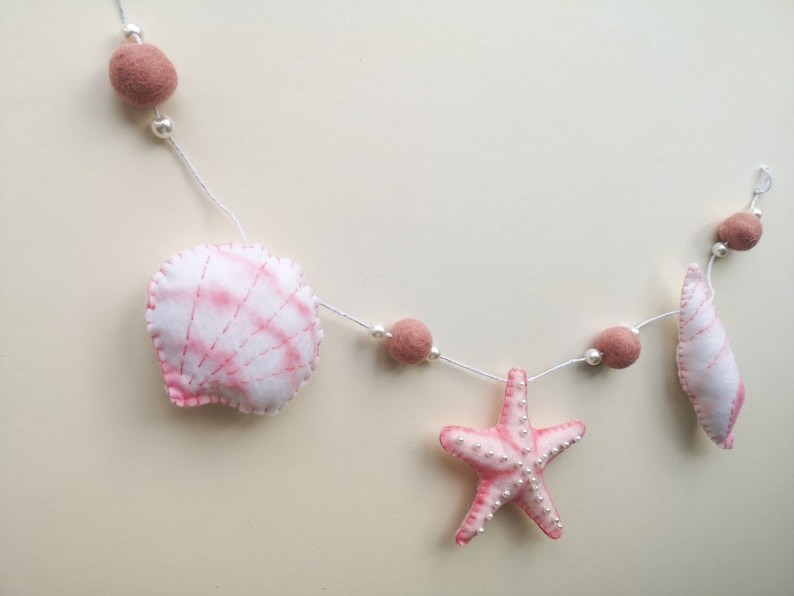 May include: A decorative garland featuring felted shapes including a white and pink scallop shell, a pink starfish, and a smaller white and pink shell. The shapes are strung on a white cord with dusty rose felt balls and small white beads.