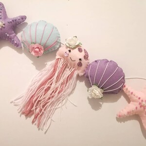 May include: A garland featuring a purple starfish, a light blue seashell, a pink jellyfish with white flowers, a purple seashell with white flowers, and a pink starfish with pink beads. The garland is perfect for a nursery or child's room.