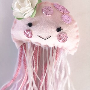 May include: A pink felt jellyfish with a white felt flower on top. The jellyfish has pink glitter dots and pink yarn tentacles.