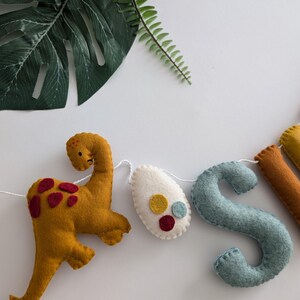 Pode incluir: Uma faixa de dinossauro de feltro com as letras "JOSH" em azul, amarelo e branco. A faixa tamb&eacute;m inclui um dinossauro amarelo e um ovo branco com bolinhas vermelhas e amarelas.