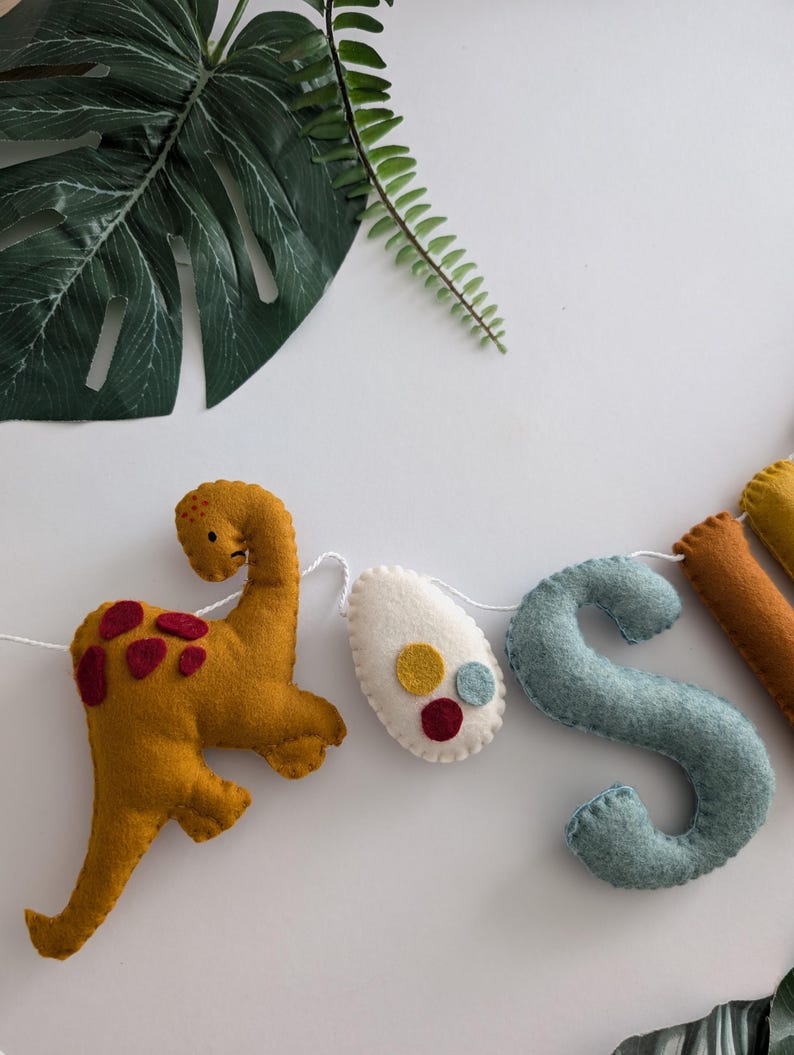 Pode incluir: Uma faixa de dinossauro de feltro com as letras "JOSH" em azul, amarelo e branco. A faixa tamb&eacute;m inclui um dinossauro amarelo e um ovo branco com bolinhas vermelhas e amarelas.