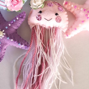 May include: A pink felt jellyfish with a white flower on top and pink and white yarn for tentacles. The jellyfish has a smiling face with black eyes and pink glitter accents.