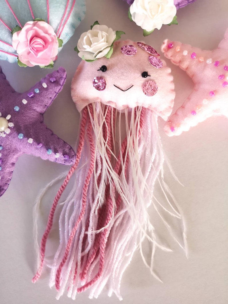 May include: A pink felt jellyfish with a white flower on top and pink and white yarn for tentacles. The jellyfish has a smiling face with black eyes and pink glitter accents.