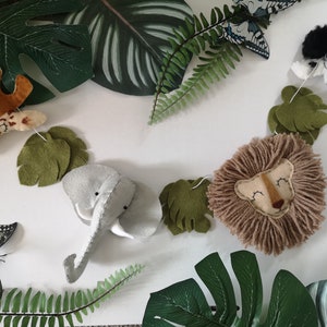 Può includere: Una ghirlanda decorativa con teste di animali in feltro tra cui una giraffa, un elefante, un leone con criniera in filato e una zebra, intervallate da foglie in feltro verde, disposte tra varie foglie tropicali artificiali e farfalle.