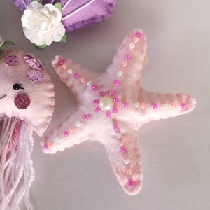May include: A pink felt starfish with pink and white beads sewn around the edges. The starfish has a pearl in the center.