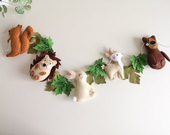 May include: A felt garland with woodland animals, including a squirrel, hedgehog, bunny, deer, and bear. The animals are brown, tan, white, and green. The garland is strung with green felt leaves.