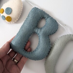Pode incluir: Uma faixa de feltro com as letras "BO" em azul e verde. As letras s&atilde;o costuradas juntas com fio branco.