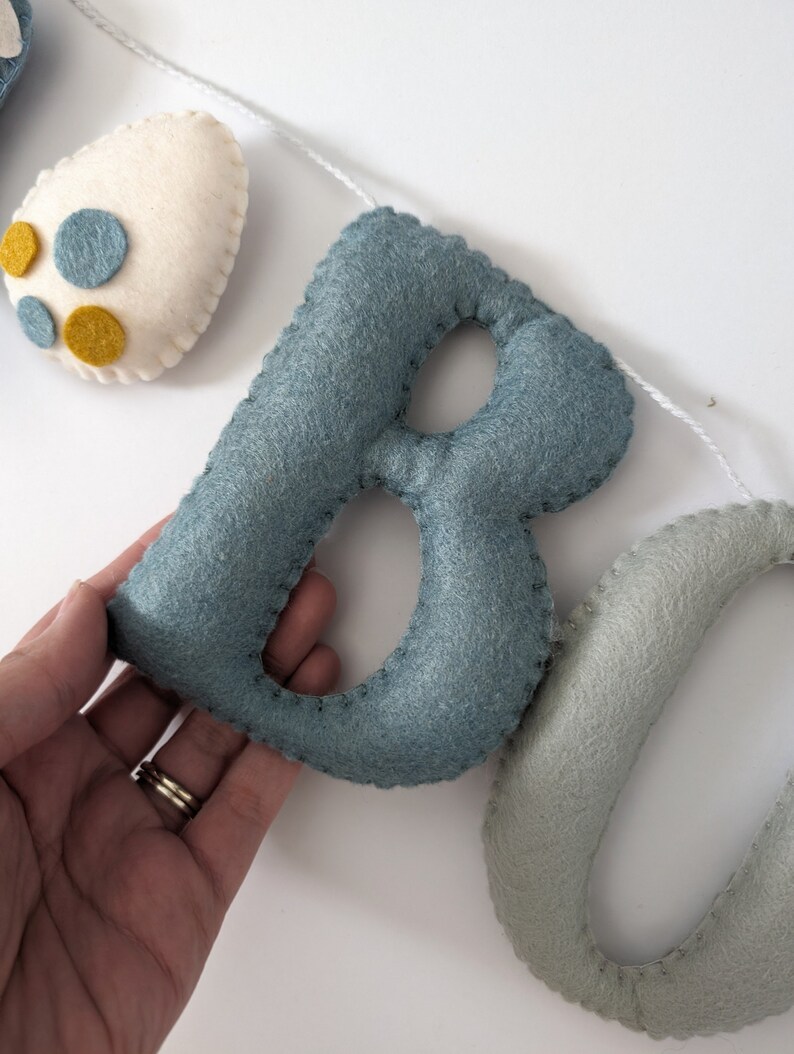 Pode incluir: Uma faixa de feltro com as letras "BO" em azul e verde. As letras s&atilde;o costuradas juntas com fio branco.
