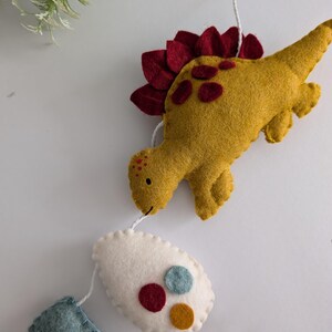 Pode incluir: Uma guirlanda de dinossauro de feltro com um dinossauro amarelo, um ovo branco com c&iacute;rculos de feltro vermelho, azul e amarelo, e um ret&acirc;ngulo azul de feltro.