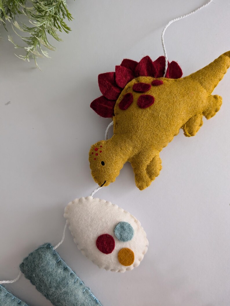 Pode incluir: Uma guirlanda de dinossauro de feltro com um dinossauro amarelo, um ovo branco com c&iacute;rculos de feltro vermelho, azul e amarelo, e um ret&acirc;ngulo azul de feltro.