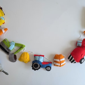 May include: A colorful felt construction-themed garland featuring various vehicles and safety equipment. The garland includes a yellow dump truck, orange traffic cones, a green excavator, a yellow hard hat, a blue tractor, and a red truck.