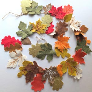 May include: A decorative garland featuring an assortment of felt leaves in autumn colours including red, orange, yellow, brown, olive green, and gold glitter. The leaves are strung together on a thin brown twine, showcasing various shapes.
