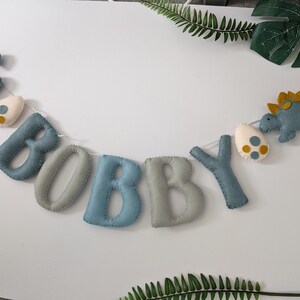 Pode incluir: Uma faixa de feltro com o nome "BOBBY" escrito em letras azuis e cinzas. A faixa &eacute; decorada com dinossauros e ovos de feltro.