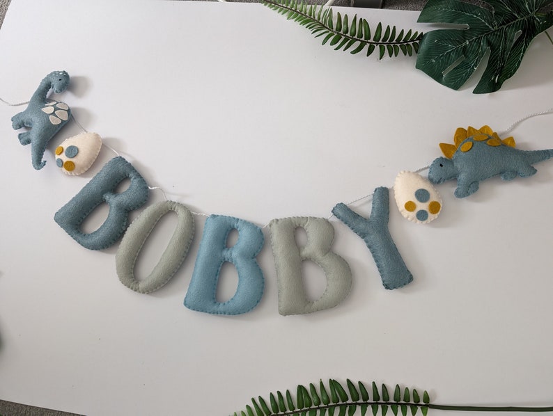Pode incluir: Uma faixa de feltro com o nome "BOBBY" escrito em letras azuis e cinzas. A faixa &eacute; decorada com dinossauros e ovos de feltro.