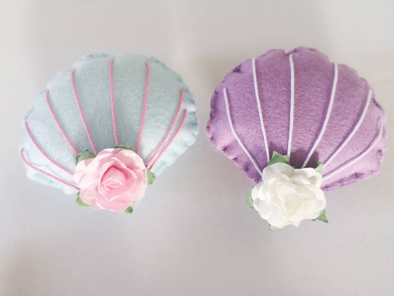 May include: Two felt seashells with pink and white fabric flowers. The seashells are light blue and purple.