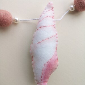 May include: A white and pink felt seashell ornament hanging on a white string. The seashell features stitched detailing and is flanked by two round, dusty pink felt balls and two small white beads on either side of the central shell piece.