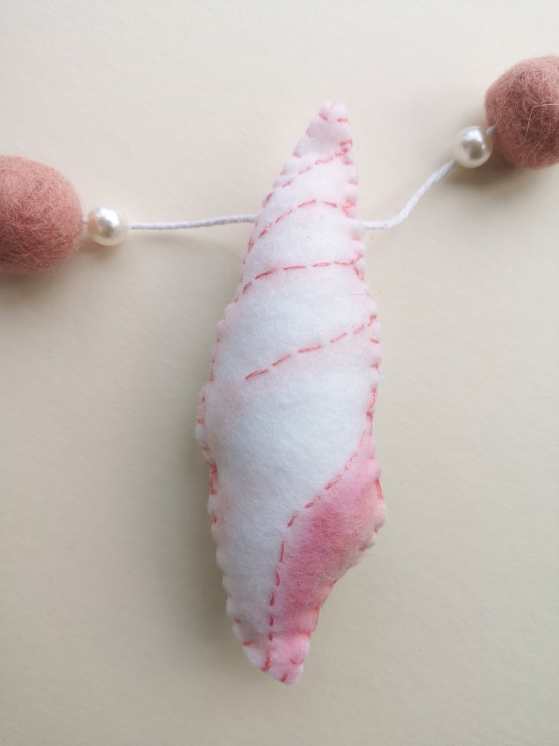 May include: A white and pink felt seashell ornament hanging on a white string. The seashell features stitched detailing and is flanked by two round, dusty pink felt balls and two small white beads on either side of the central shell piece.