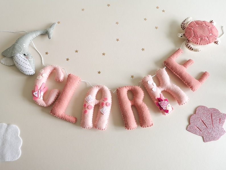 May include: A pink felt banner with the name "CLARE" spelled out in individual letters. Each letter is decorated with small pink and white felt embellishments. A grey felt whale and a pink felt turtle are attached to the banner.