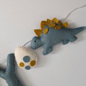 Pode incluir: Uma faixa de dinossauro de feltro com um dinossauro azul, um ovo branco com manchas amarelas e uma letra Y azul. O dinossauro tem uma crista espinhosa amarela e manchas amarelas.