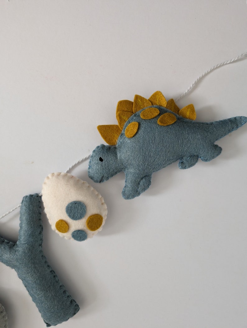 Pode incluir: Uma faixa de dinossauro de feltro com um dinossauro azul, um ovo branco com manchas amarelas e uma letra Y azul. O dinossauro tem uma crista espinhosa amarela e manchas amarelas.