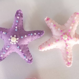 May include: Two felt starfish ornaments, one purple and one pink, with pearl-like beads and white beads sewn around the edges.