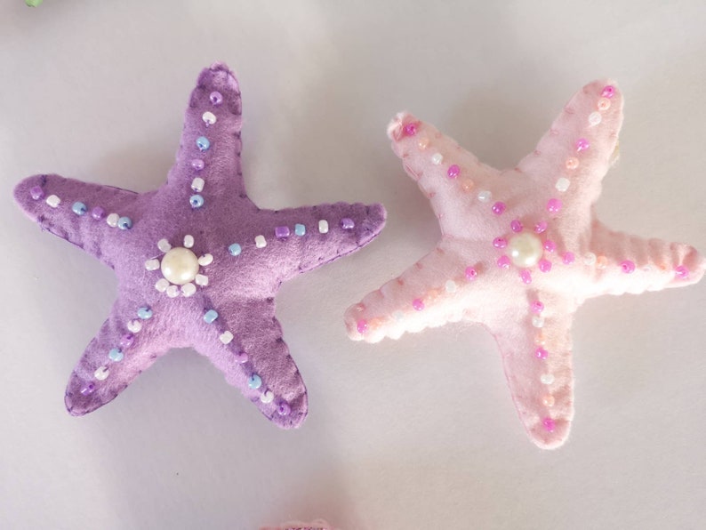 May include: Two felt starfish ornaments, one purple and one pink, with pearl-like beads and white beads sewn around the edges.