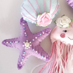 May include: A set of felt ocean-themed ornaments, including a purple starfish with white and purple beads, a light blue seashell with pink stitching and a pink flower, and a pink jellyfish with white and pink yarn tentacles.