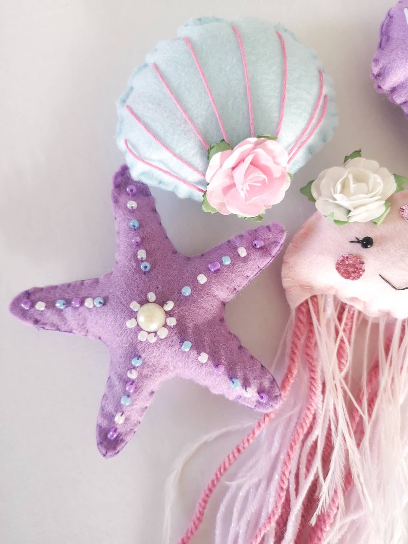 May include: A set of felt ocean-themed ornaments, including a purple starfish with white and purple beads, a light blue seashell with pink stitching and a pink flower, and a pink jellyfish with white and pink yarn tentacles.