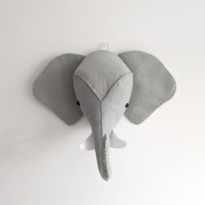 May include: A grey felt elephant head wall decoration with large ears, black eyes, and a white tusk. The elephant head is mounted on a white wall, suitable for a nursery or children's room.