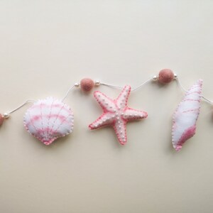 May include: A decorative garland featuring soft felt shapes including a scallop shell, a starfish, and a conch shell, all in shades of white and pink. The items are strung on a white cord interspersed with small white beads and dusty pink felt balls.