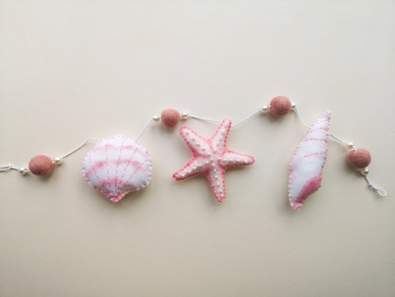 May include: A decorative garland featuring soft felt shapes including a scallop shell, a starfish, and a conch shell, all in shades of white and pink. The items are strung on a white cord interspersed with small white beads and dusty pink felt balls.